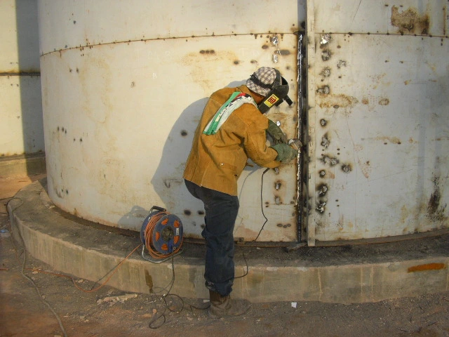 Welder sedang melakukan pengelasan pada sambungan vertikal dinding tangki.